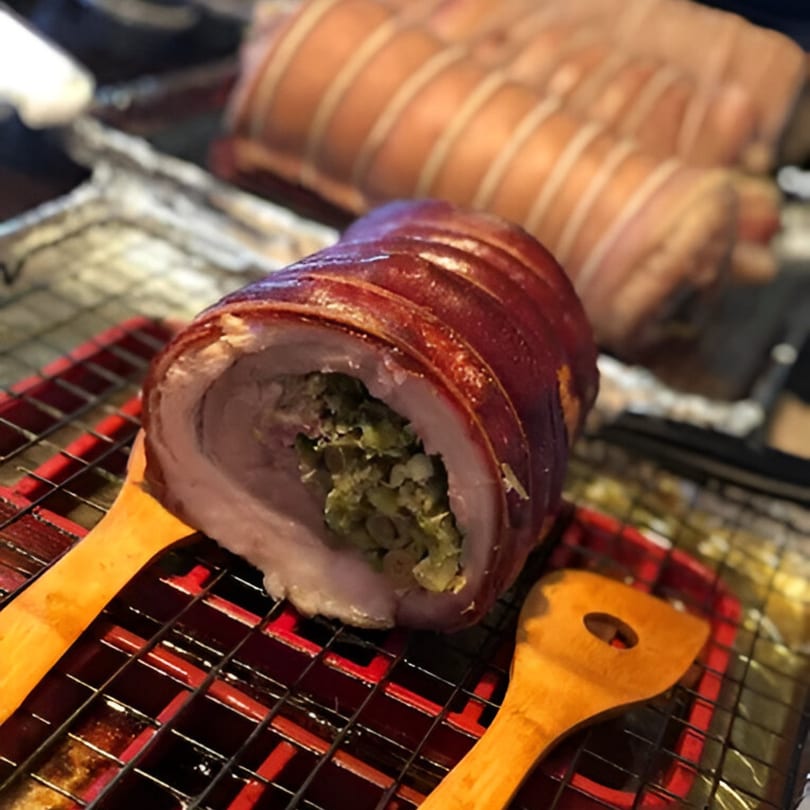 A large, stuffed pork belly roll with crispy skin rests on a red and black grill rack.
