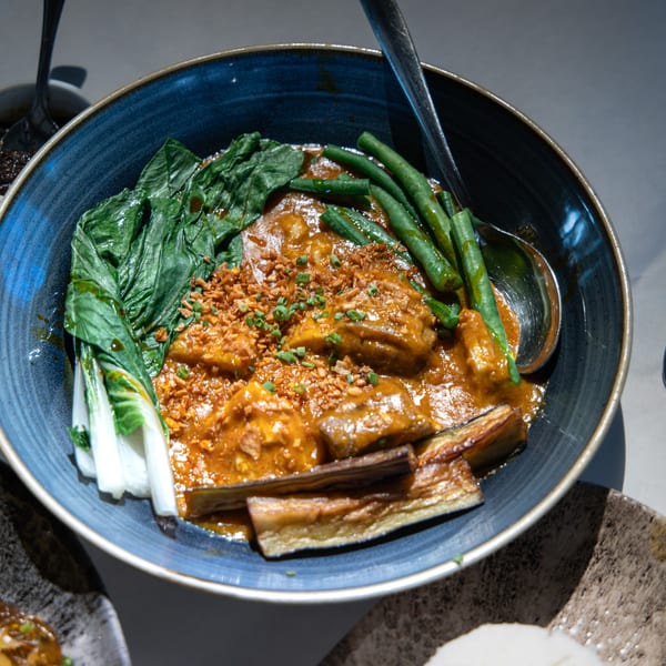 A blue bowl of kare-kare with bok choy, green beans, eggplant, and a spoon.