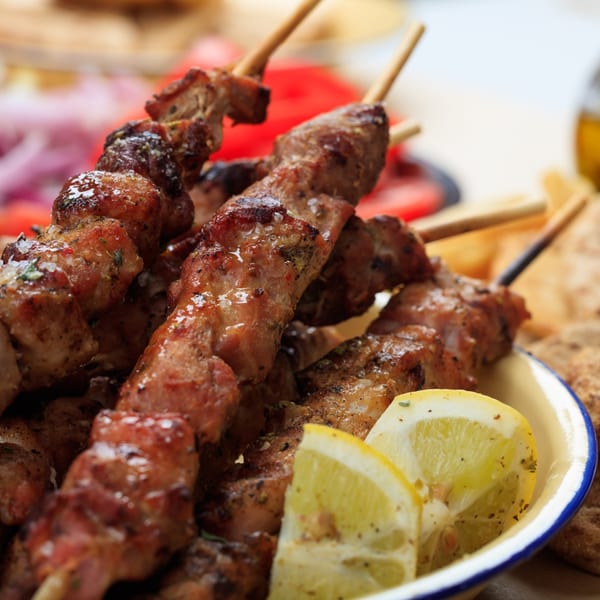 Grilled meat skewers with lemon slices in a white bowl, with red onions and tomatoes in the background.