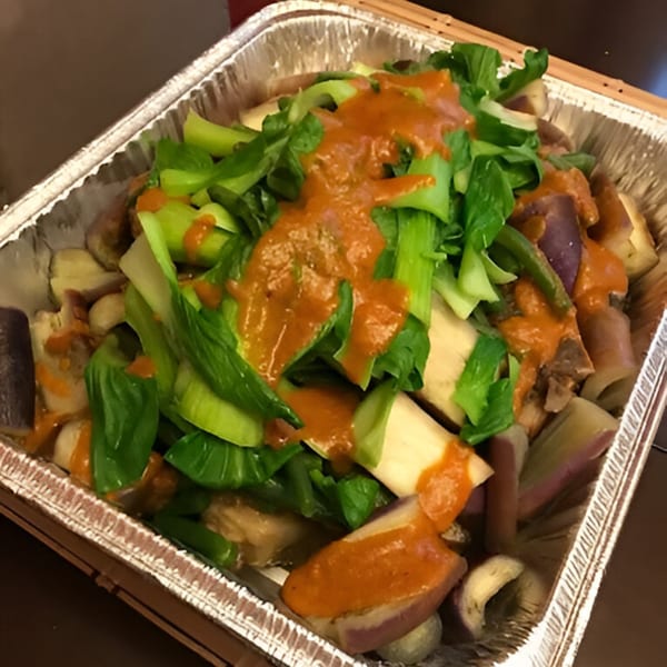 A foil tray filled with green vegetables, purple eggplant, and other ingredients, topped with orange sauce.
